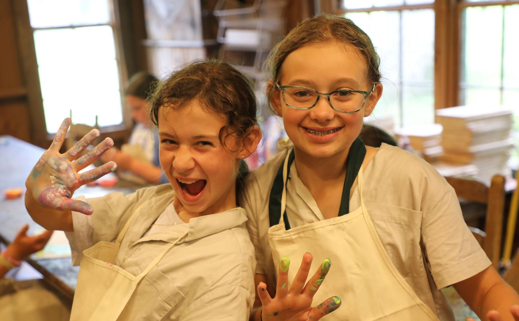 Two children in white aprons showing paint-covered hands and smiling in a workshop setting.