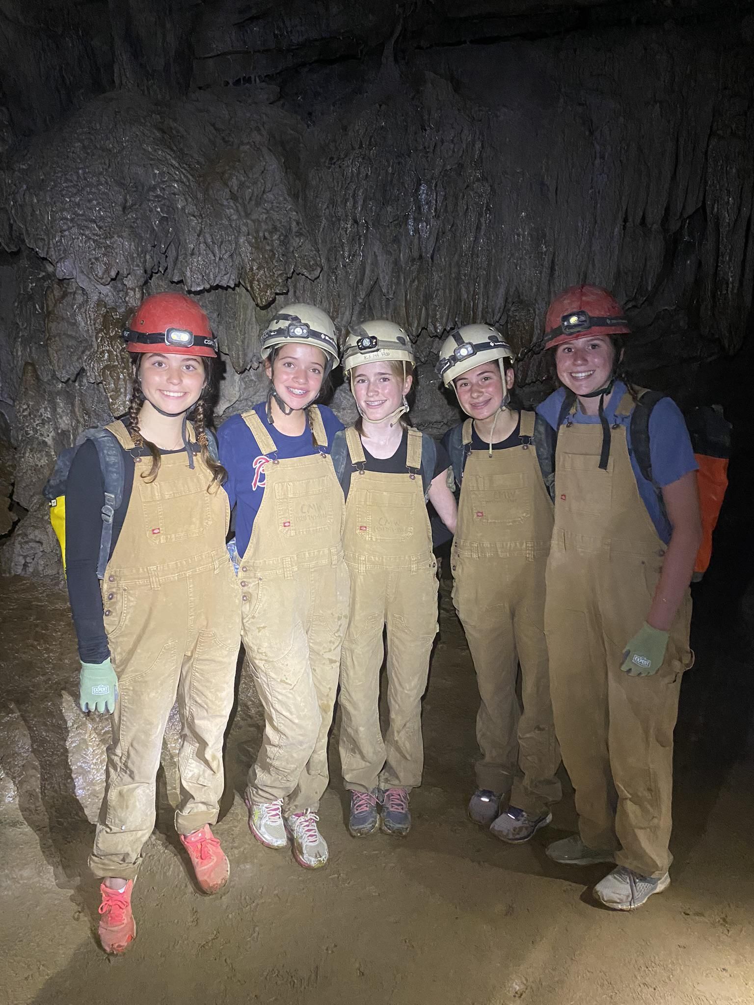 Five people wearing helmets and tan coveralls stand together smiling inside a dark, rocky cave.