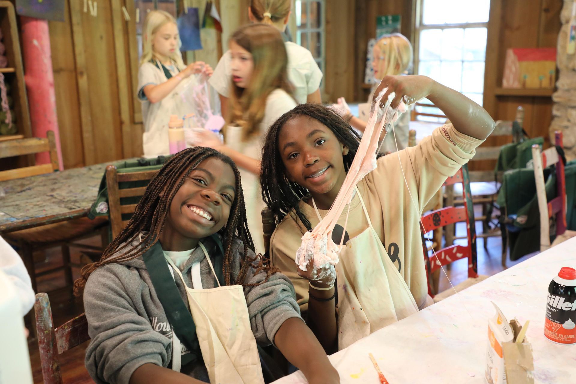 Two smiling children sit at a table in a rustic workshop, one stretching a strand of light-colored slime toward the camera.