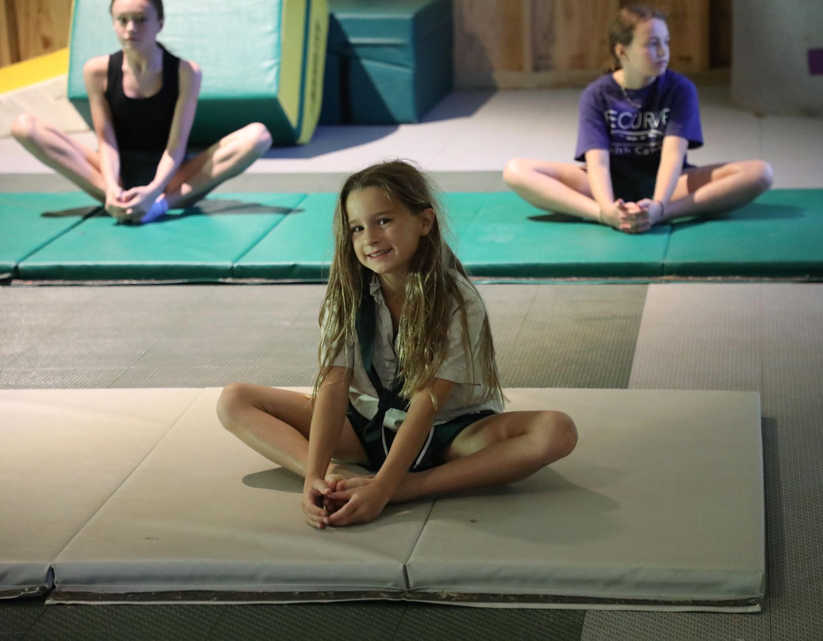 Three children sit on gymnastics mats in a butterfly stretch, with one smiling at the camera in the foreground.