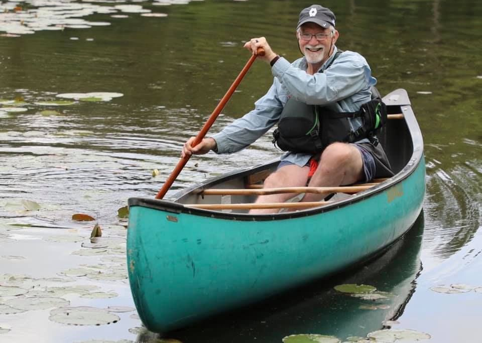 A person smiling while paddling a teal canoe on a calm, lily-pad-covered pond.