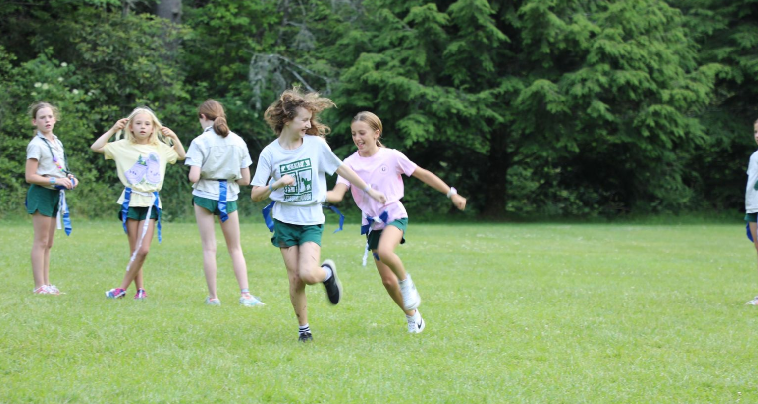Several children play flag football on a large, grassy field surrounded by trees.