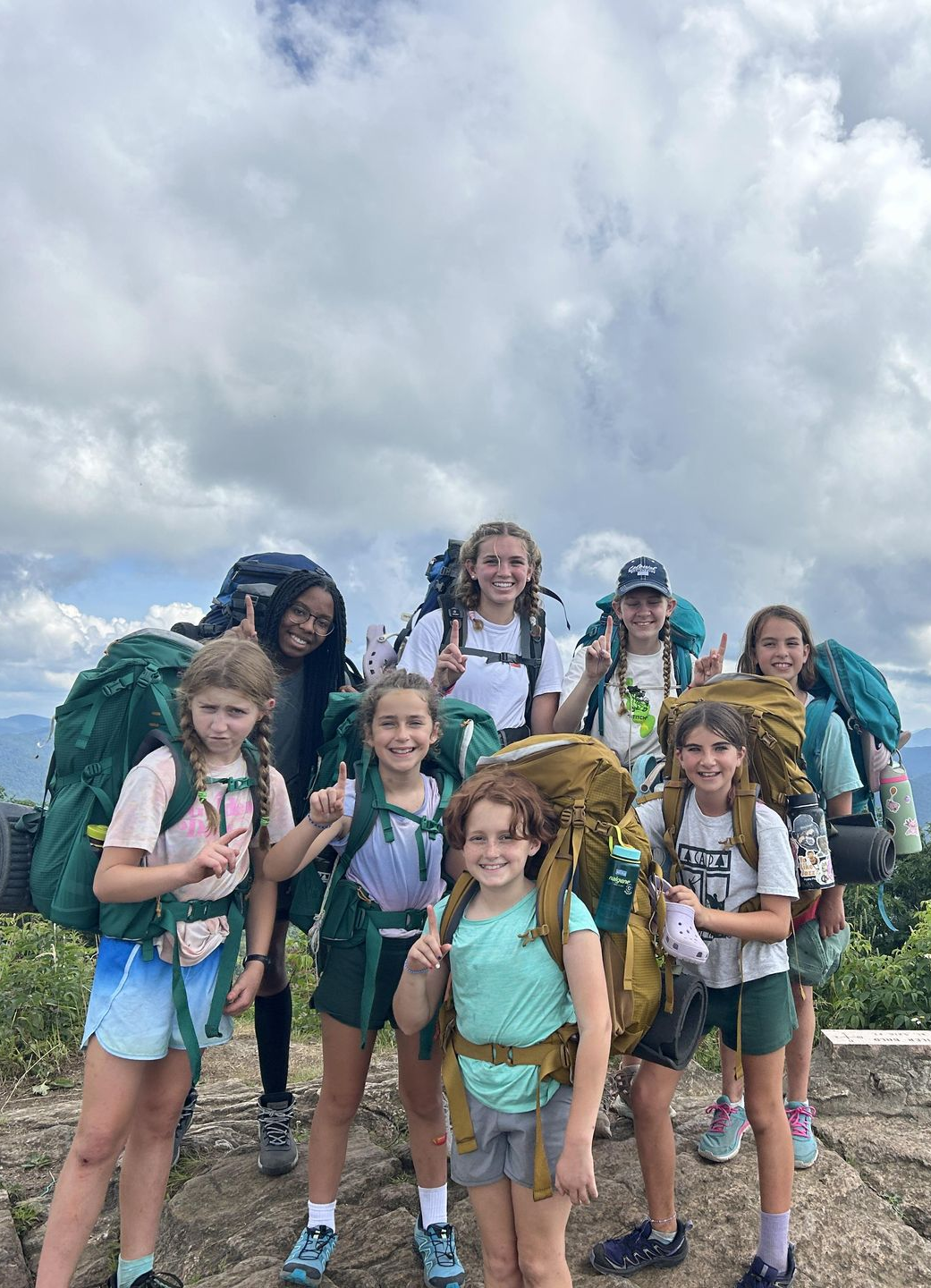 A group of hikers with large backpacks pose for a photo on a rocky mountain summit under a cloudy sky.