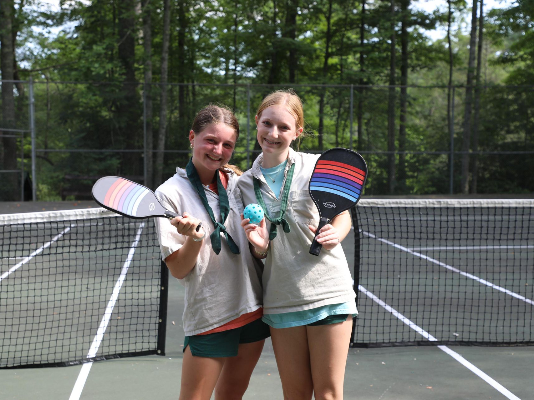 Two people standing on a pickleball court, smiling and holding matching striped paddles and a blue ball.