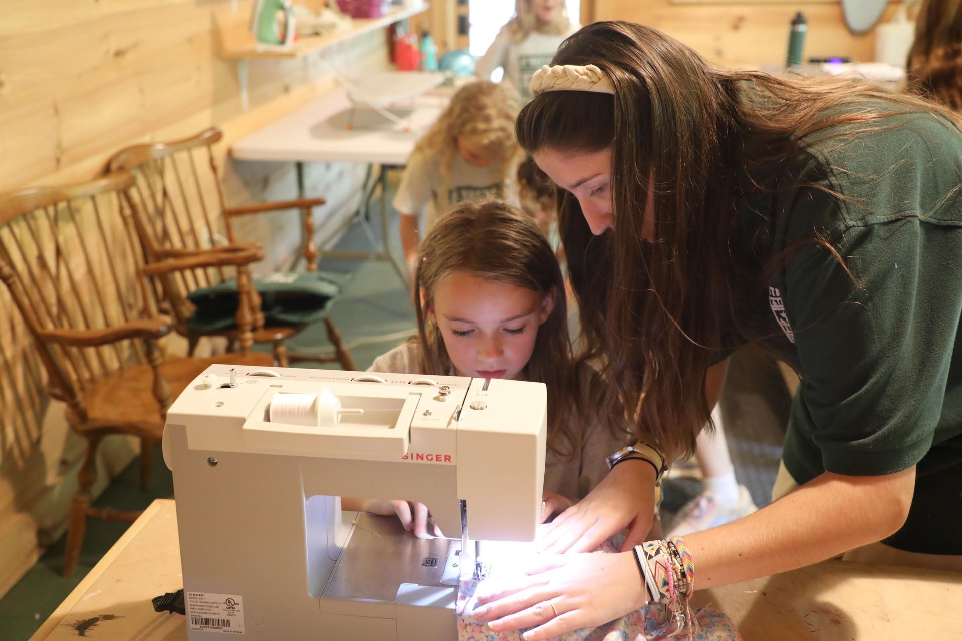 A person assists a child with a sewing machine at a table inside a wooden building.