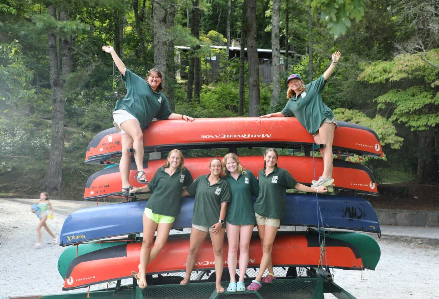 Six people in green shirts pose with a rack of stacked canoes in a wooded setting, with one person walking in the background.