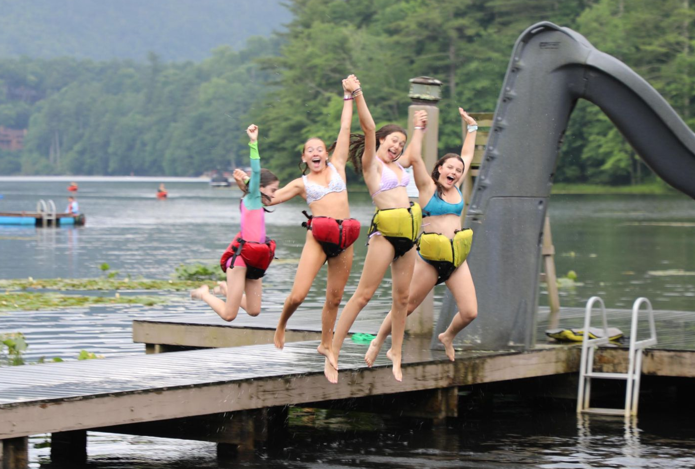 Four young people jump joyfully from a wooden dock into a lake near a slide, all wearing colorful life jackets.