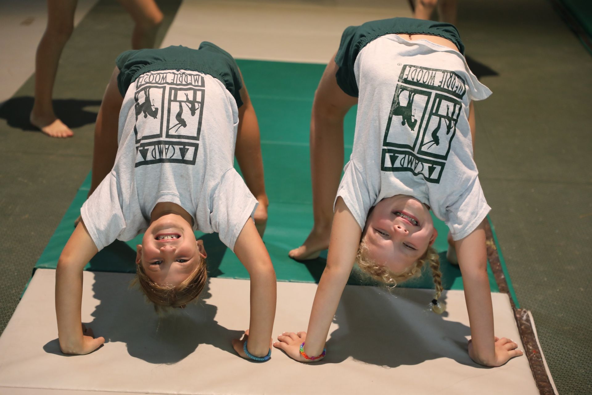 Two children perform backbends on a gymnastics mat, wearing white logo t-shirts and dark green shorts.