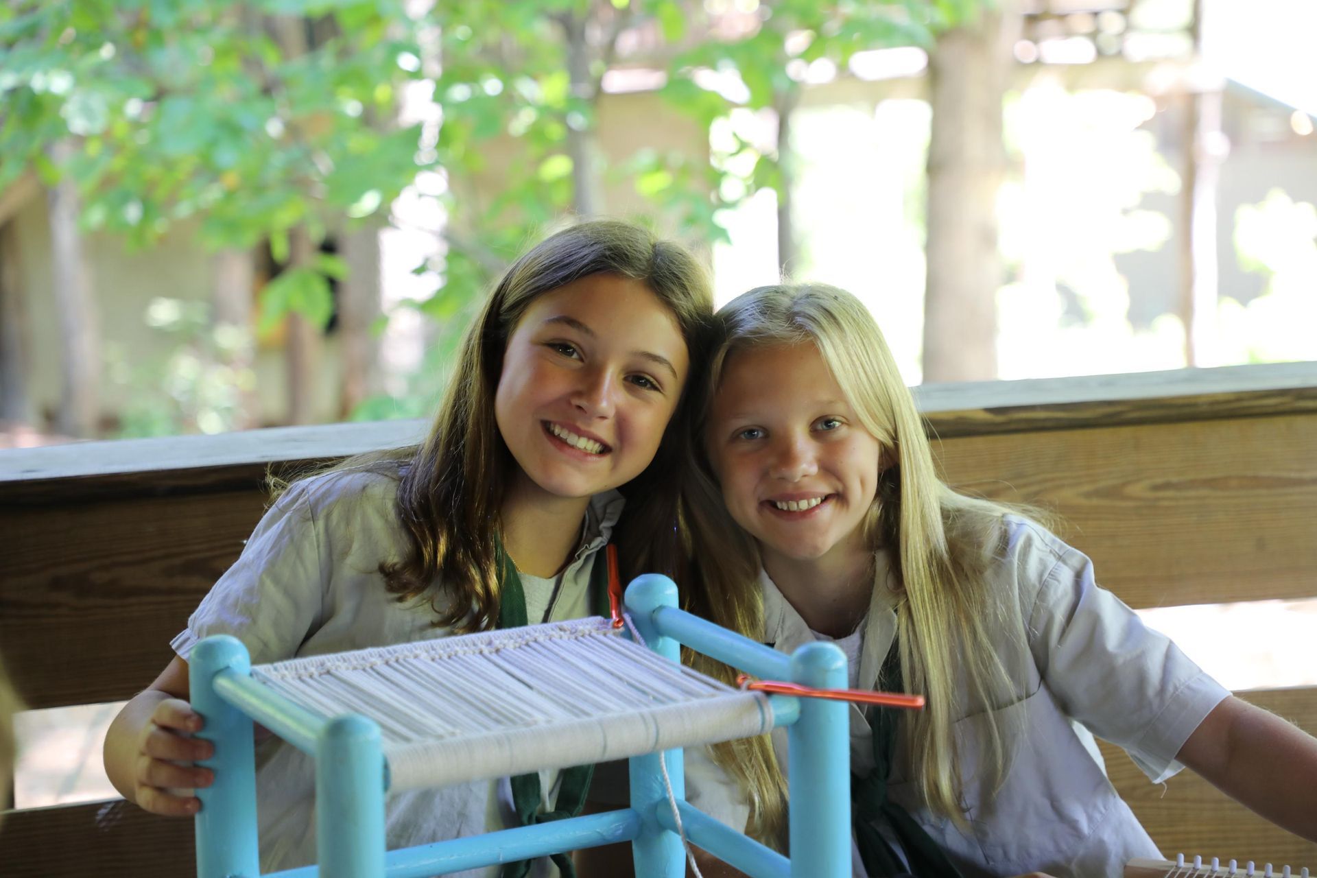 Two smiling people sit at a wooden table together, collaborating on a project involving a small, blue-framed loom.