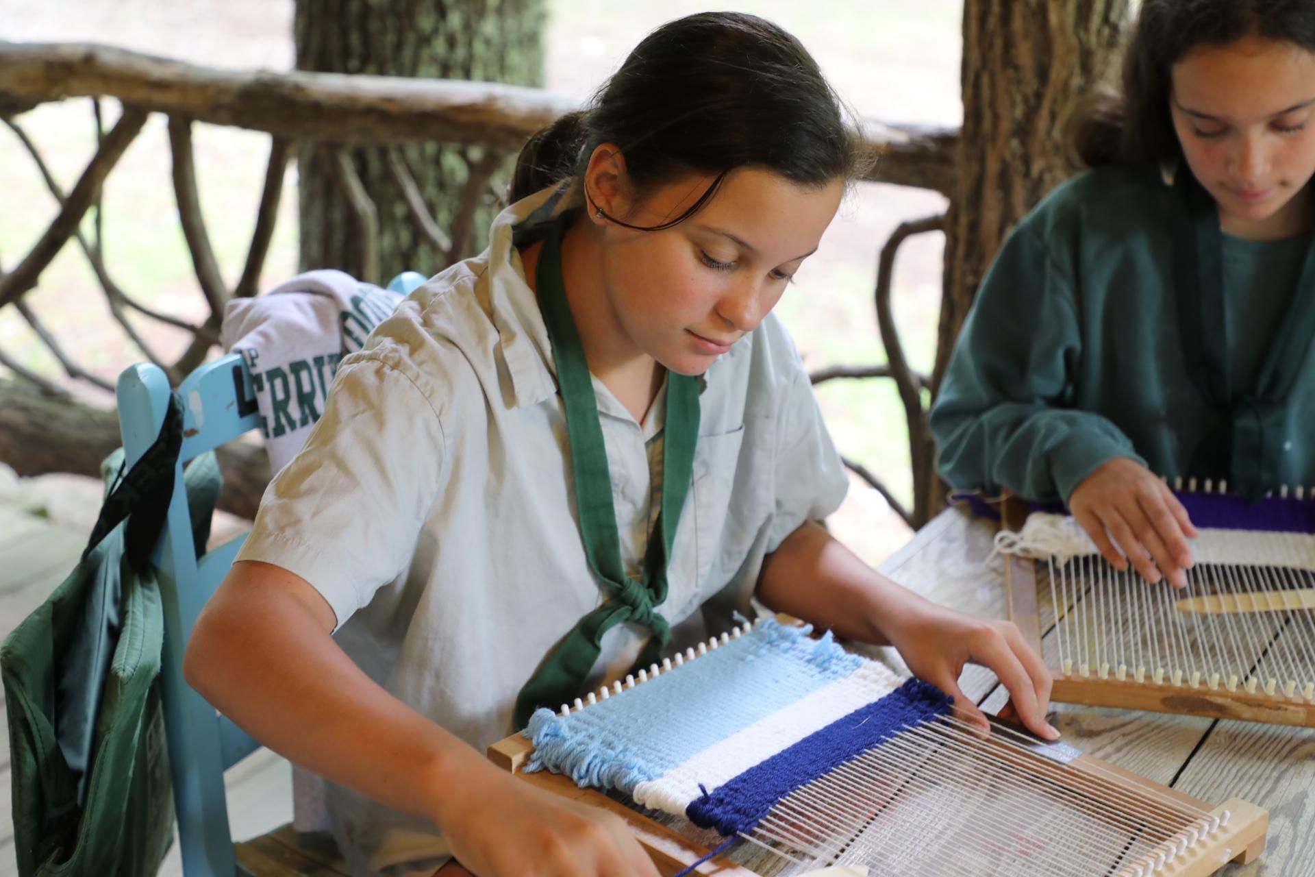 Two people focused on weaving projects on small wooden looms at a table in a rustic, outdoor wooden structure.