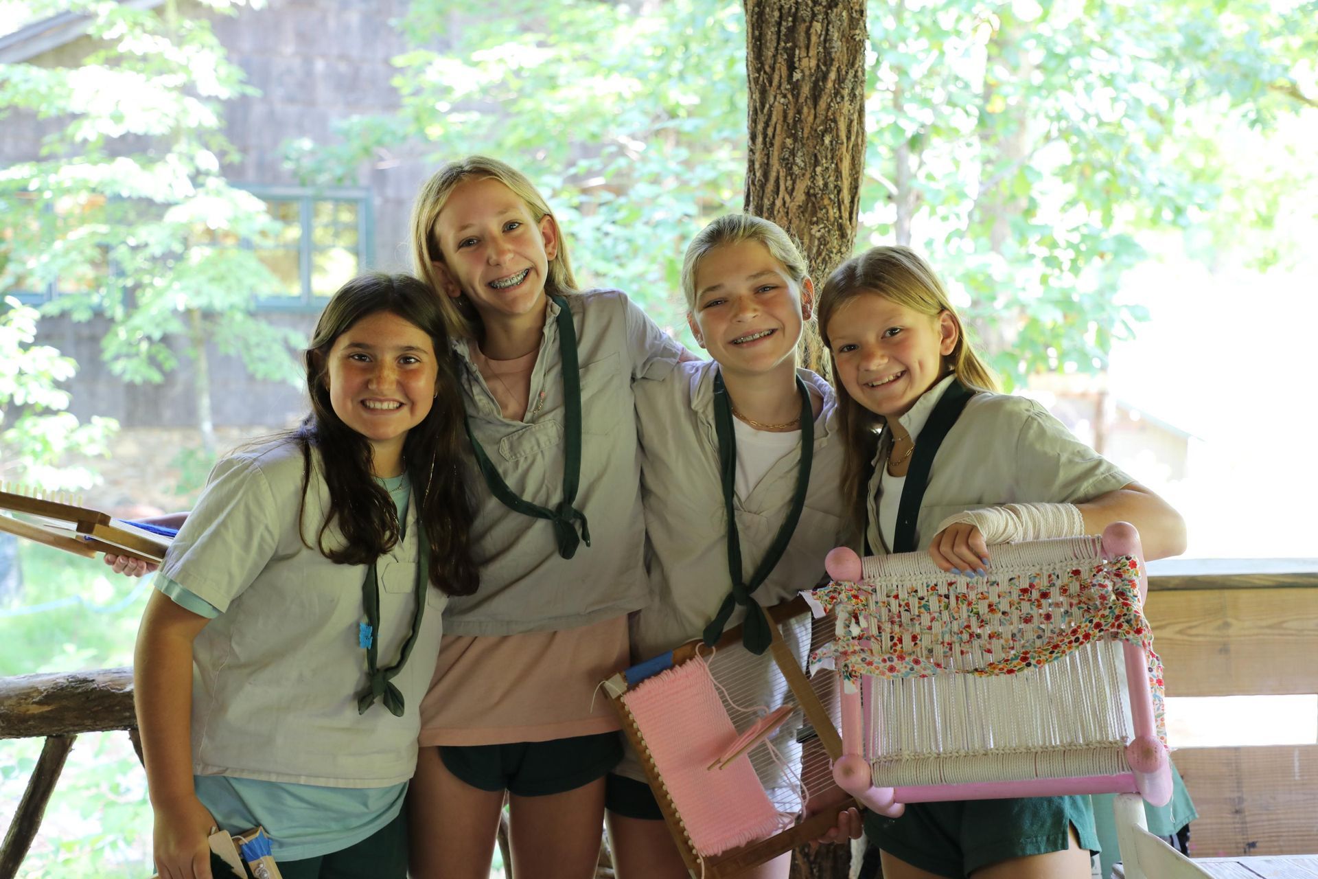 Four smiling campers in matching gray shirts and dark scarves pose outdoors, holding colorful craft projects.