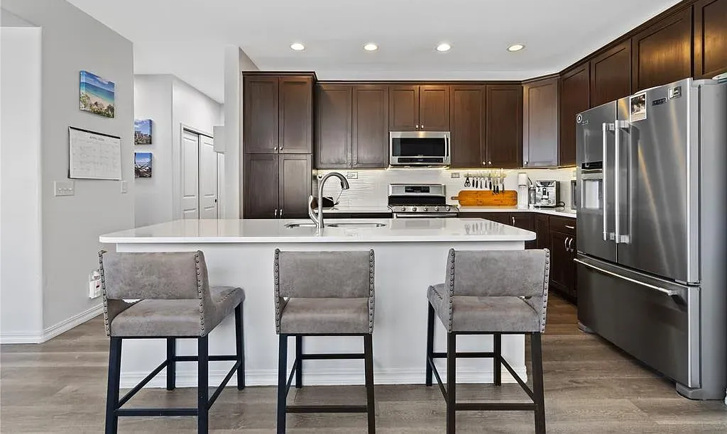 Modern kitchen with stainless steel appliances, dark cabinets, and three upholstered stools at a breakfast bar