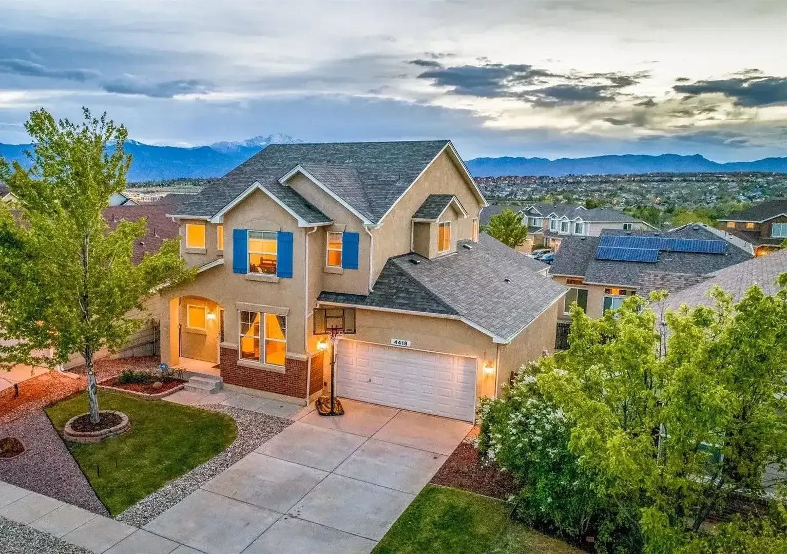Two-story suburban house with warm lights, a three-car garage, and mountain views at dusk