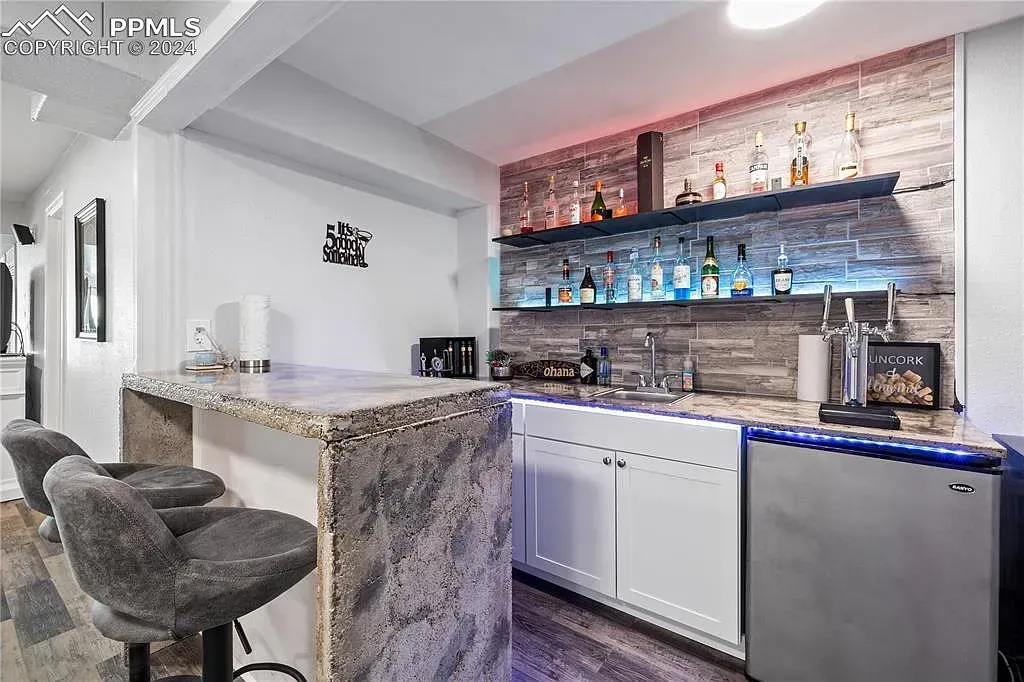 Modern home bar with granite counter, mirrored shelves, and gray upholstered stools in a bright white room