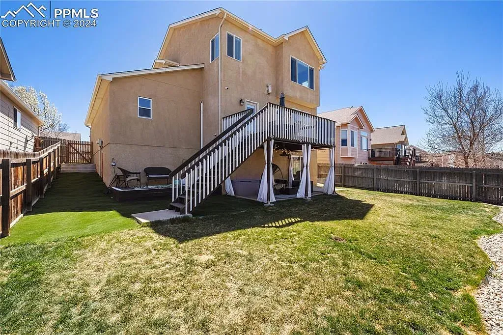 Backyard view of tan house with raised deck, black stairs, and green lawn under a blue sky