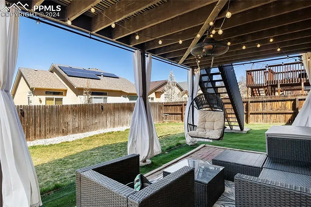 Covered backyard patio with wicker seating, hanging curtains, and string lights overlooking a fenced yard and houses.