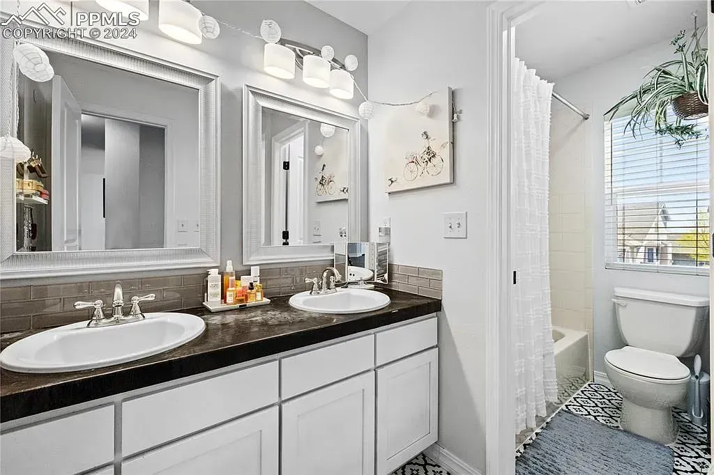 Bright white bathroom with double vanity, large mirrors, and a window beside a toilet and shower curtain.