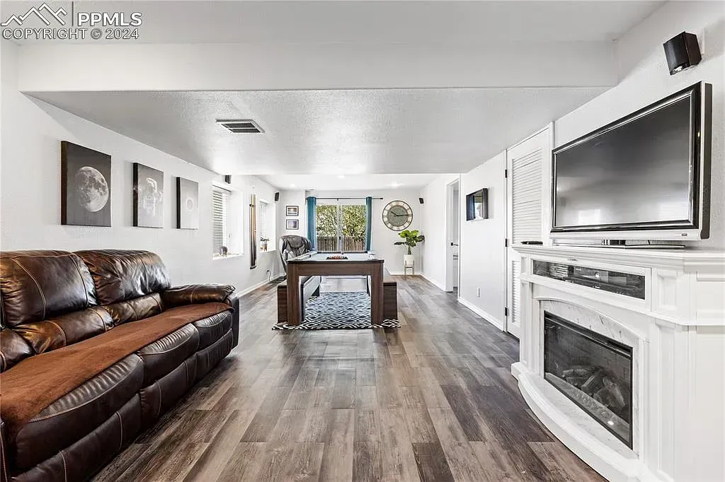Bright living room with brown leather sofa, wall-mounted TV, fireplace, and dining table with windows at the far end