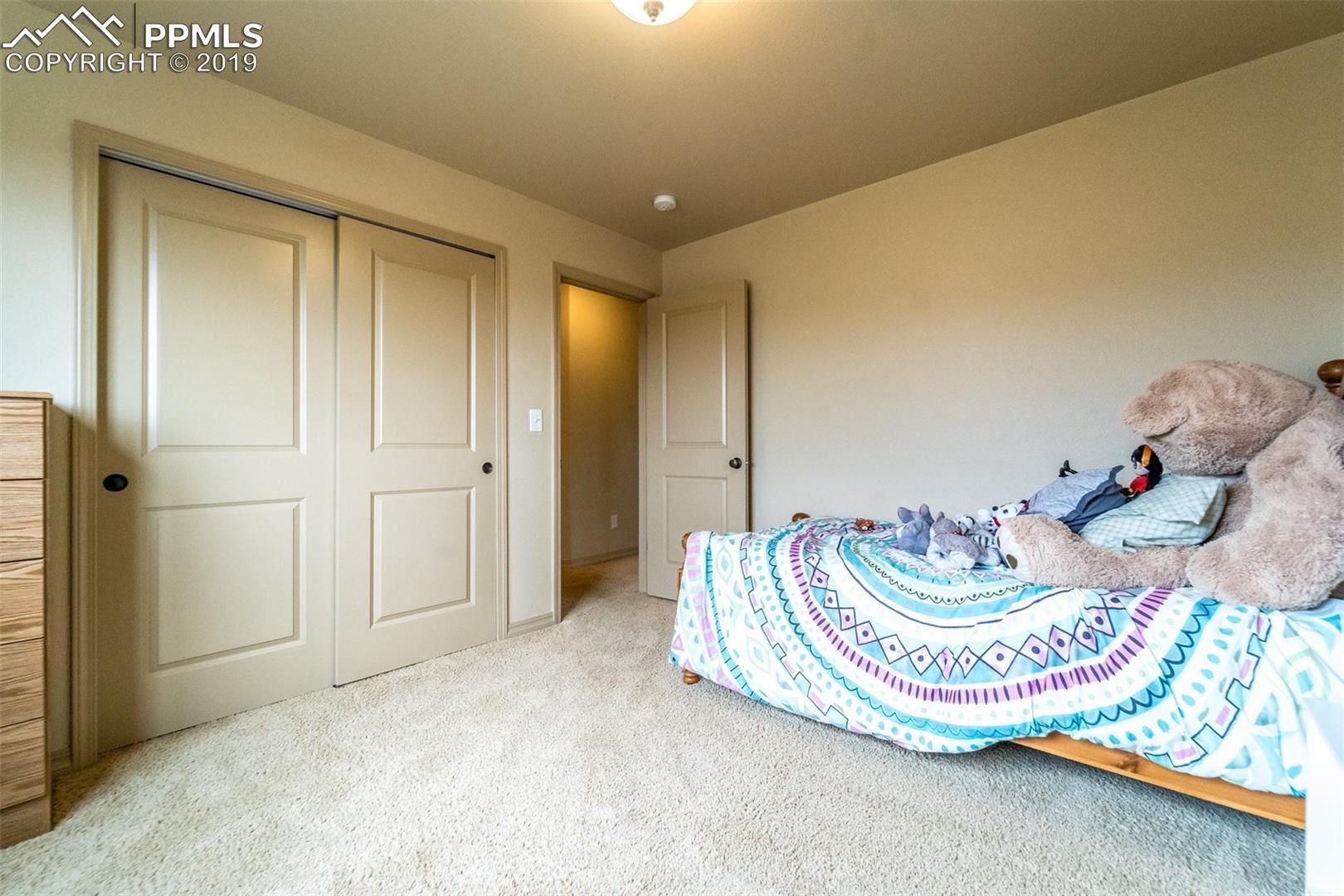 Bright bedroom with beige carpet, closet doors, and a bed covered in a colorful quilt.