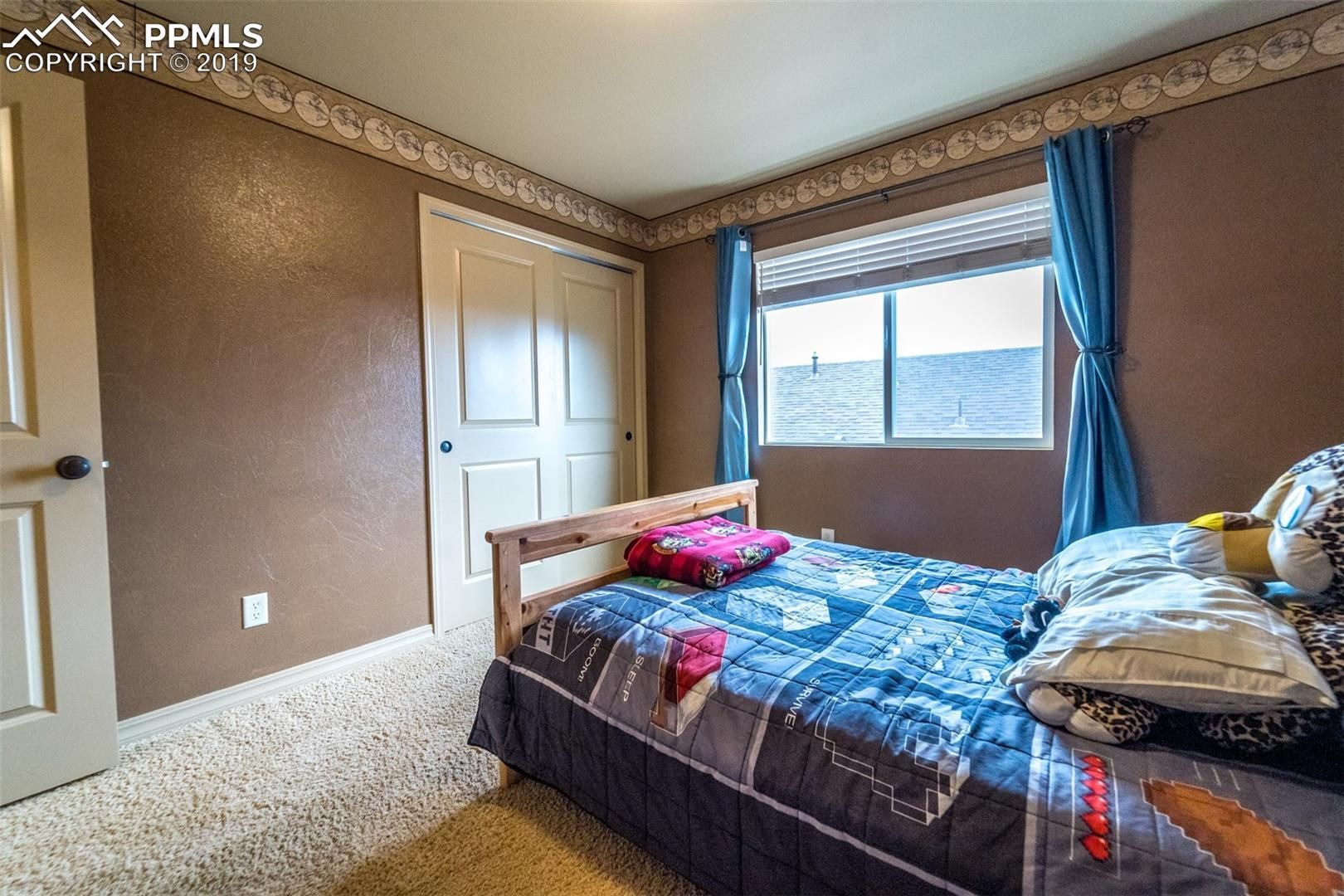 Small bedroom with a bed, blue curtains, beige walls, and a window letting in daylight