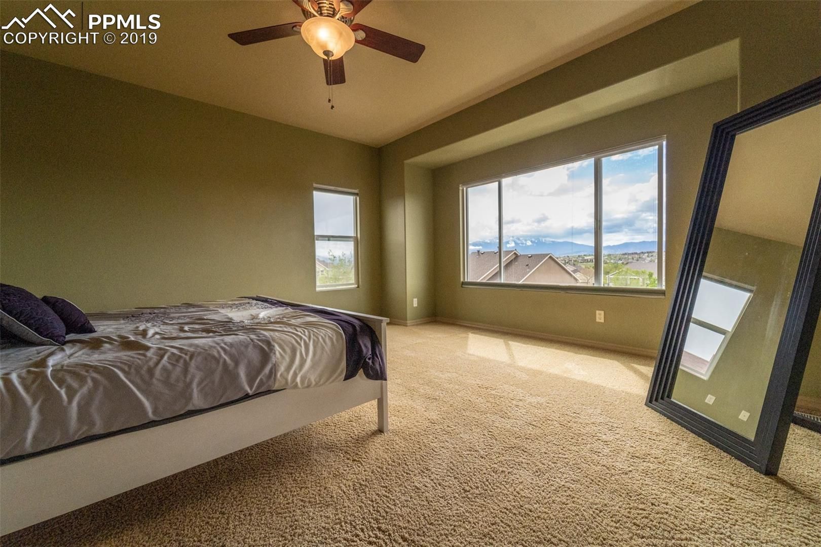 Bedroom with a bed, ceiling fan, large windows, and a full-length mirror on carpeted floor.