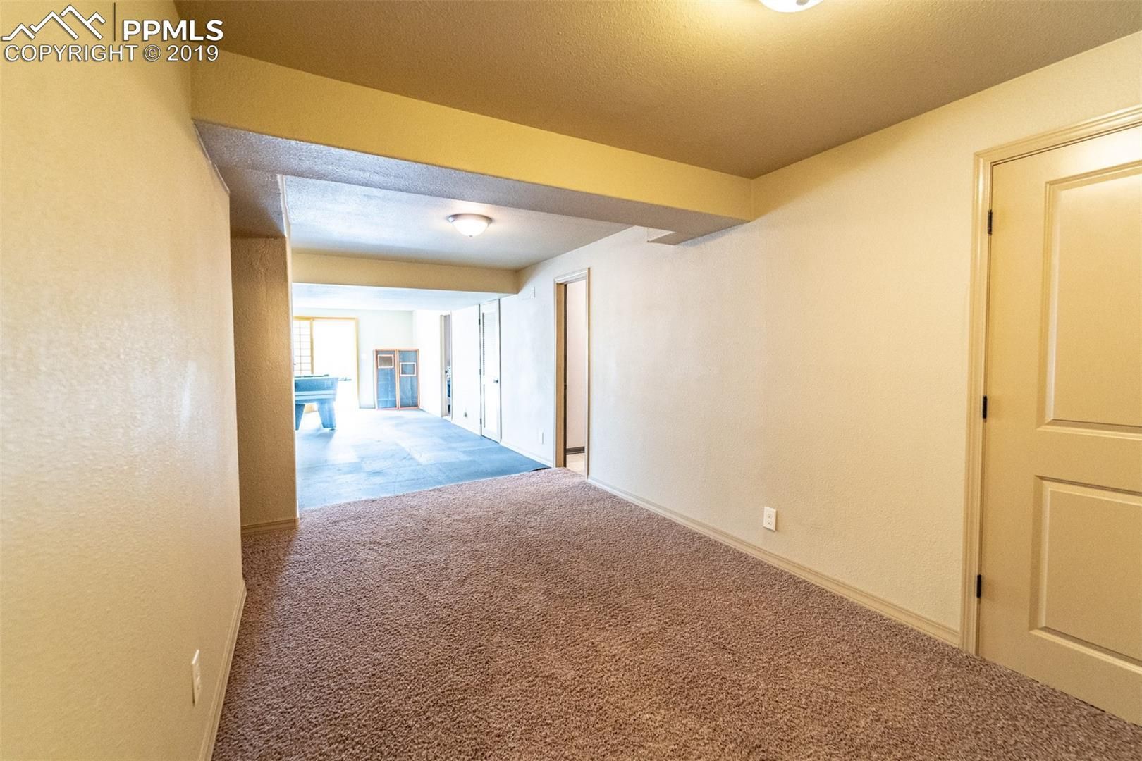 Empty carpeted hallway with beige walls, doors, and a bright living area visible at the far end.