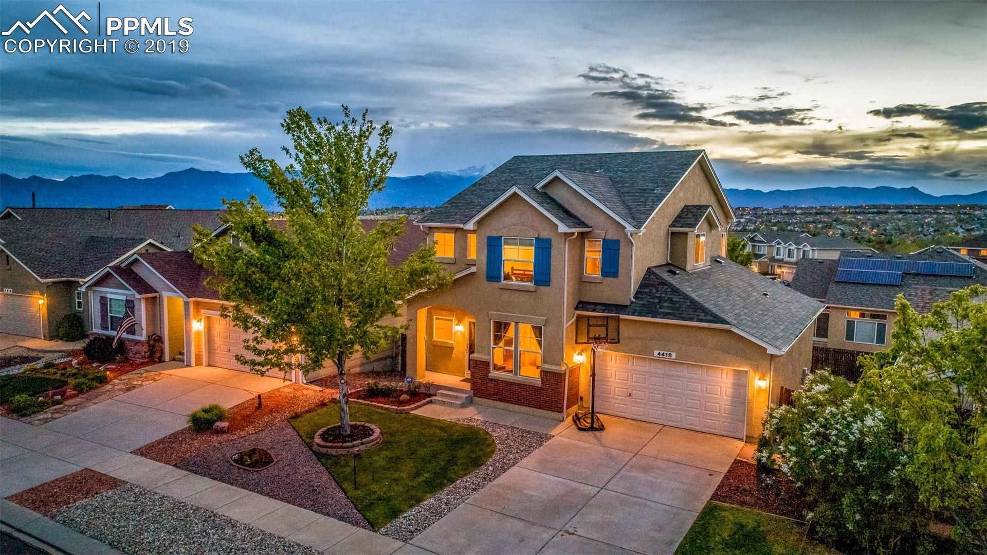 Twilight view of a modern two-story house with lit windows, driveway, and mountain backdrop