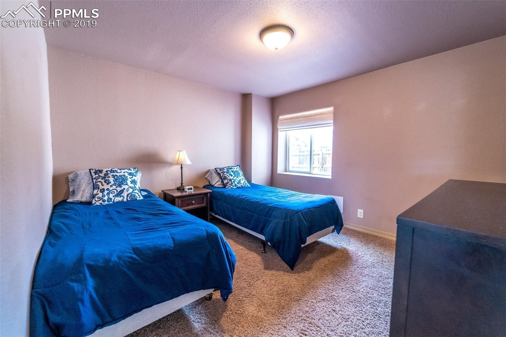 Bedroom with two twin beds, blue bedding, a window, and a dresser on carpeted floor.