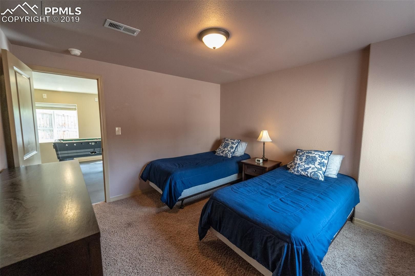 Small bedroom with two blue twin beds, nightstand lamp, and open doorway to a brighter room
