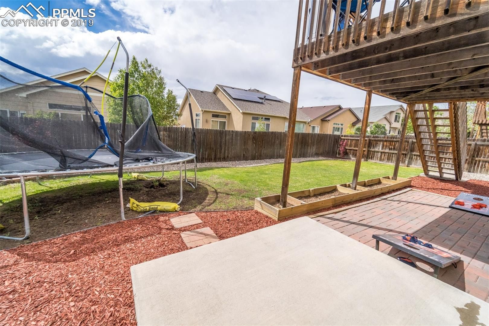 Backyard patio with trampoline, wooden deck, grass, and red gravel around a concrete slab