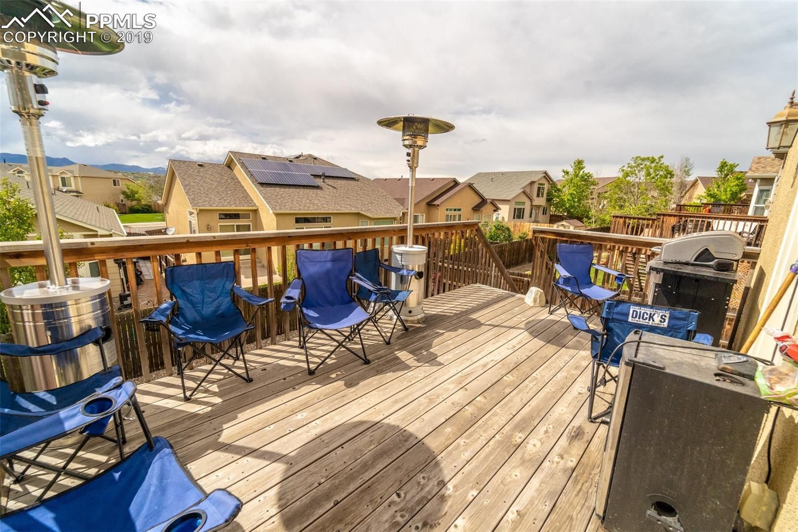Wooden rooftop deck with blue chairs, tables, and patio heaters overlooking houses under a sunny sky