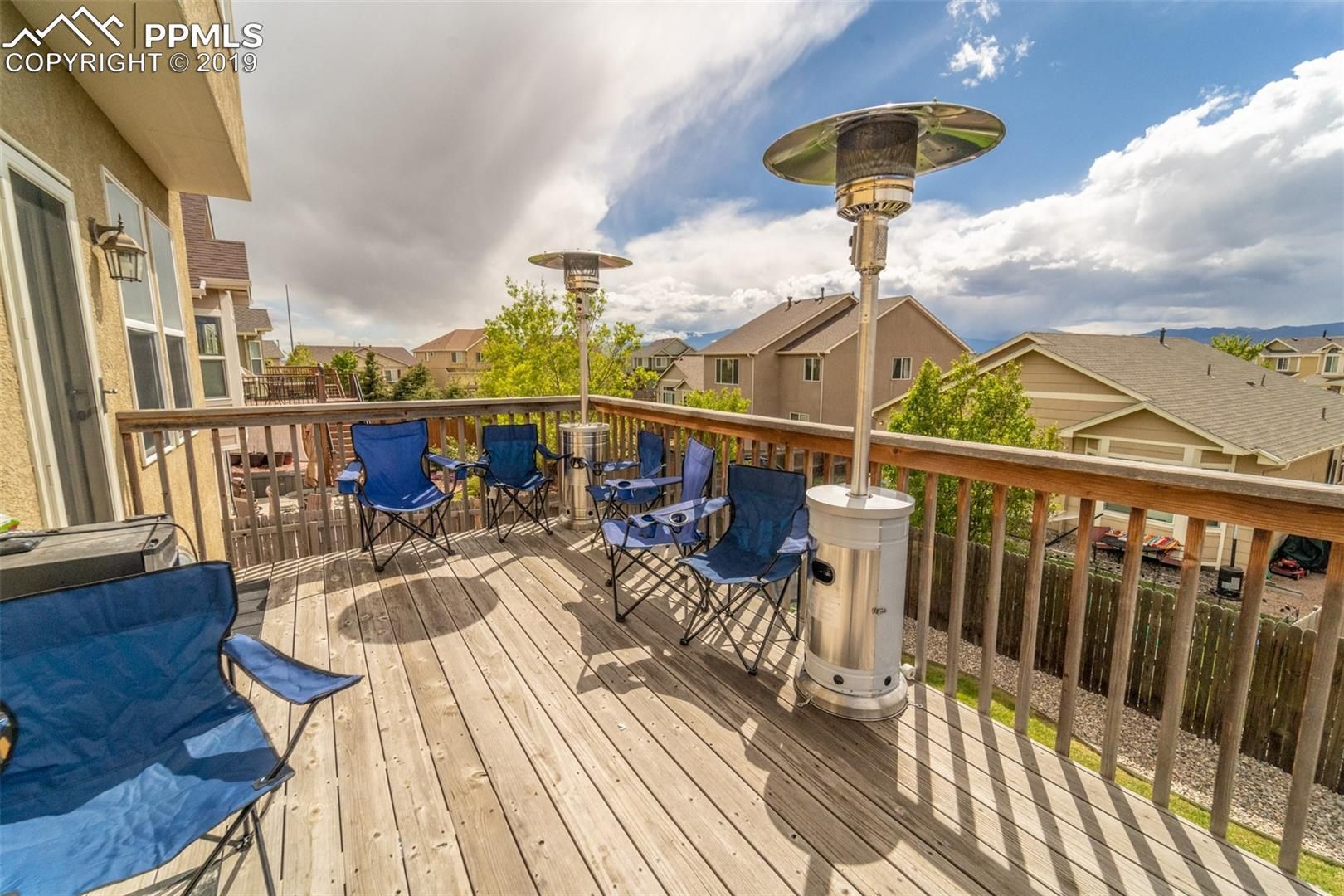 Sunlit wooden deck with blue chairs, patio heaters, railing, and neighborhood rooftops under a partly cloudy sky