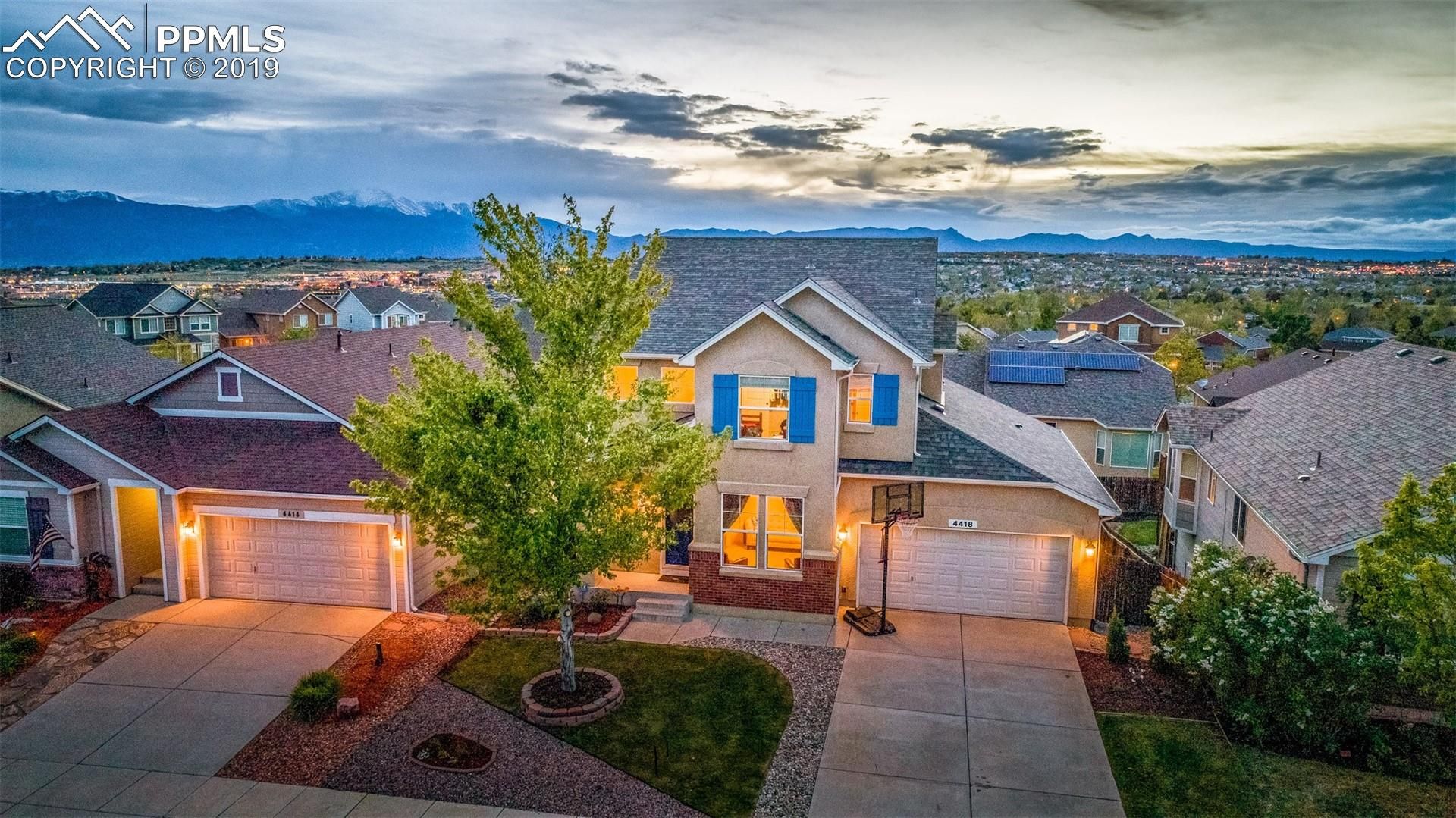 Suburban house at dusk with lit windows, driveway, and mountain views under a cloudy sky.