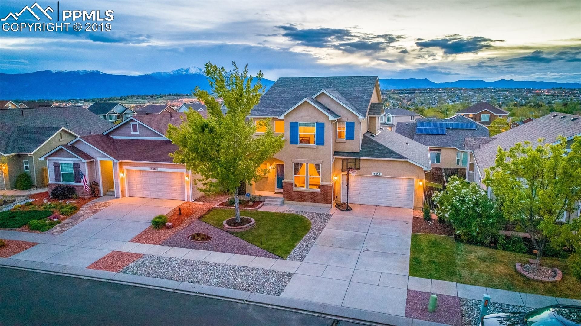 Modern two-story suburban house with stone facade, lit windows, driveway, and landscaped front yard at dusk