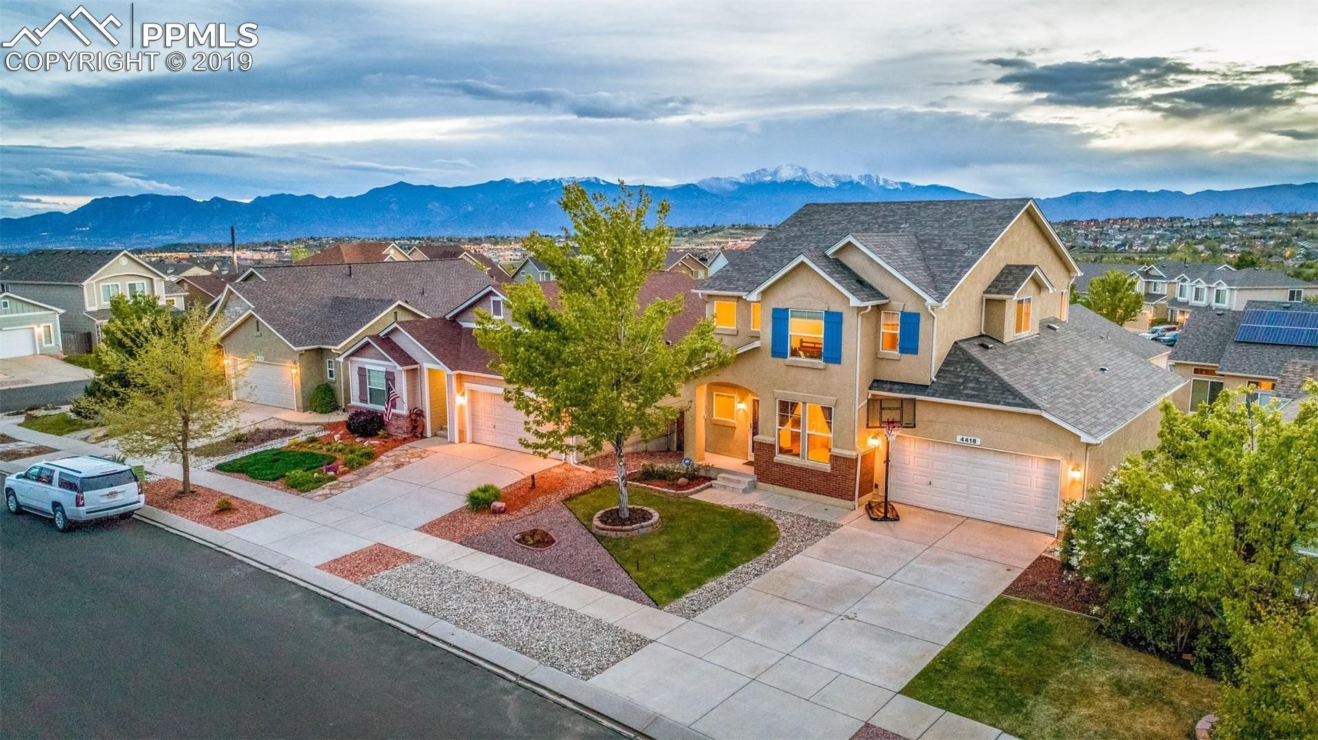 Suburban homes at dusk on a quiet street, with lit windows, parked cars, and mountain views.