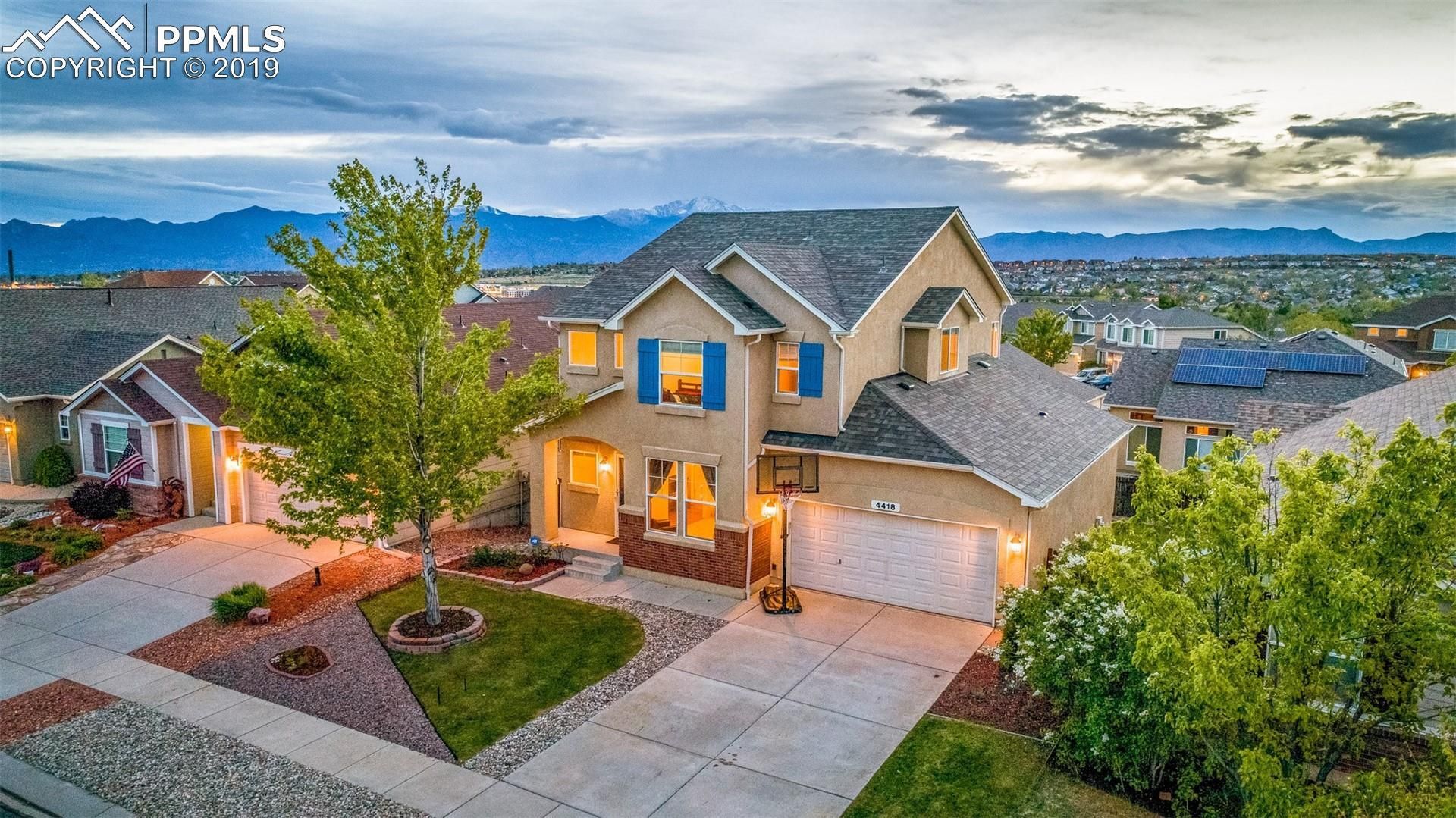 Modern two-story house with lit windows, driveway, and mountain views at dusk