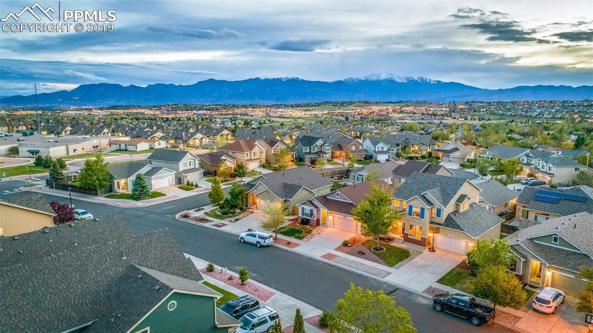 Suburban neighborhood at dusk with rows of houses, parked cars, lit streetlights, and mountains under a cloudy sky