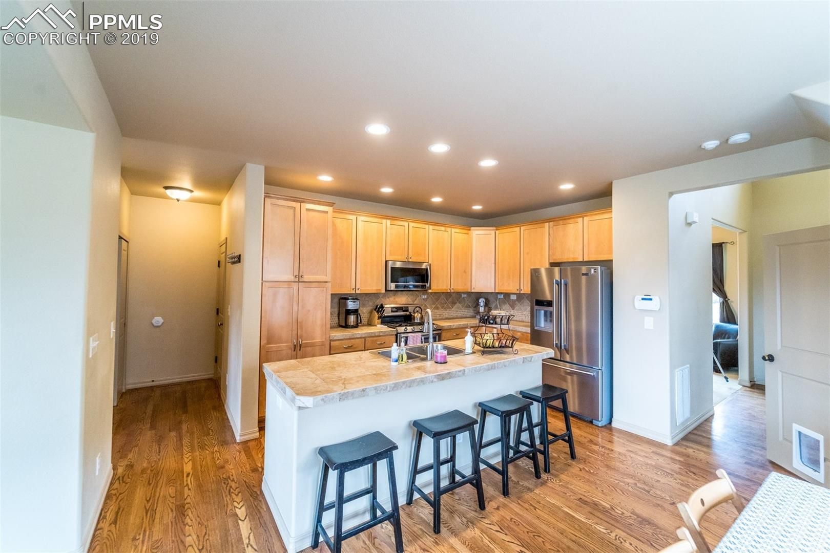 Bright kitchen with wood floors, light cabinets, granite island, and three stools; hallway and living area visible