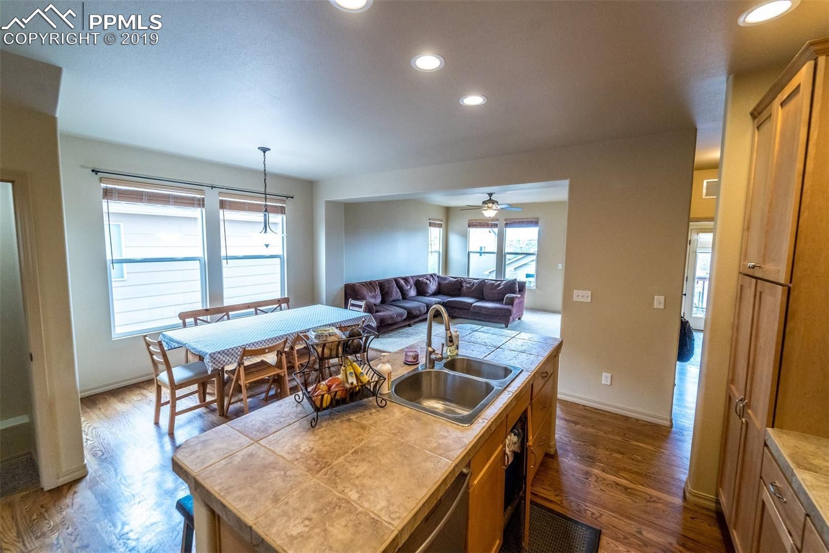 Bright open kitchen and dining area with granite island, wood floors, and large windows, leading to a living room.