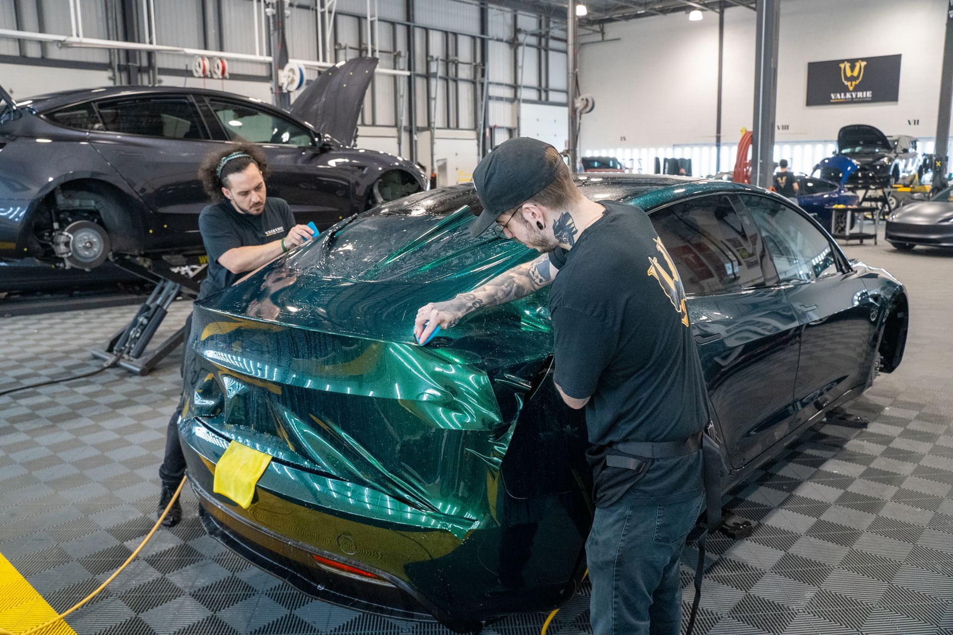 Two men are wrapping a green car in a garage.