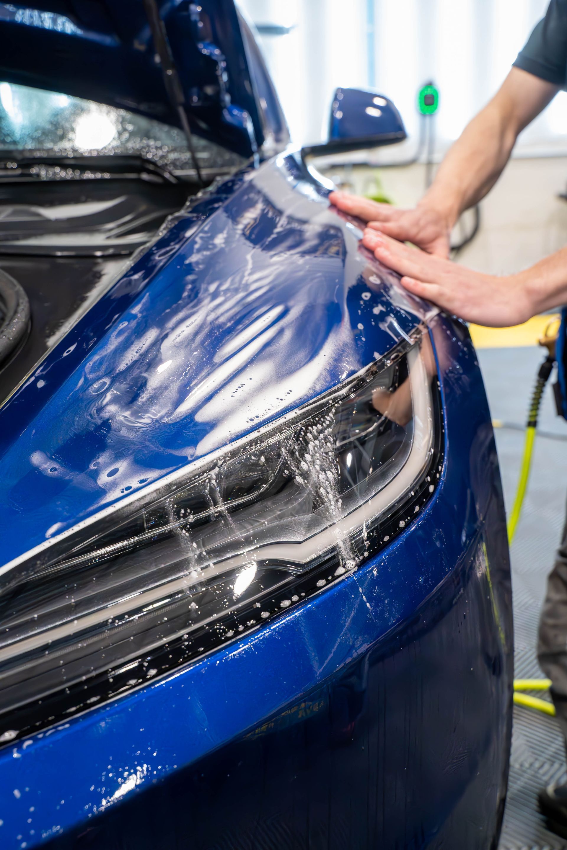 A man is applying a protective film to the headlight of a blue car.