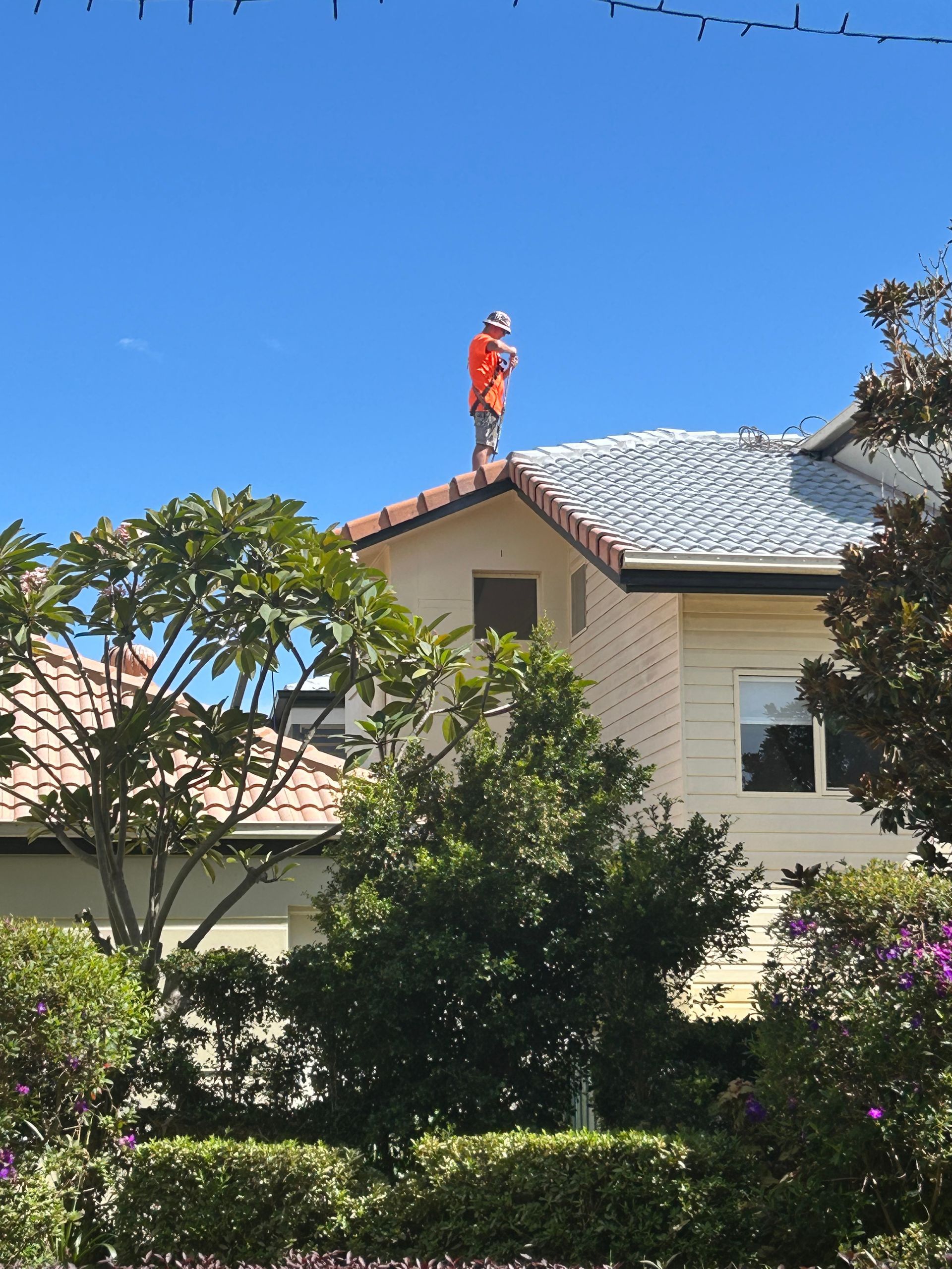 Person in orange outfit on a rooftop, surveying. Blue sky, house with trees — Magic Wand Roof Restoration & Painting in Mermaid Beach, QLD