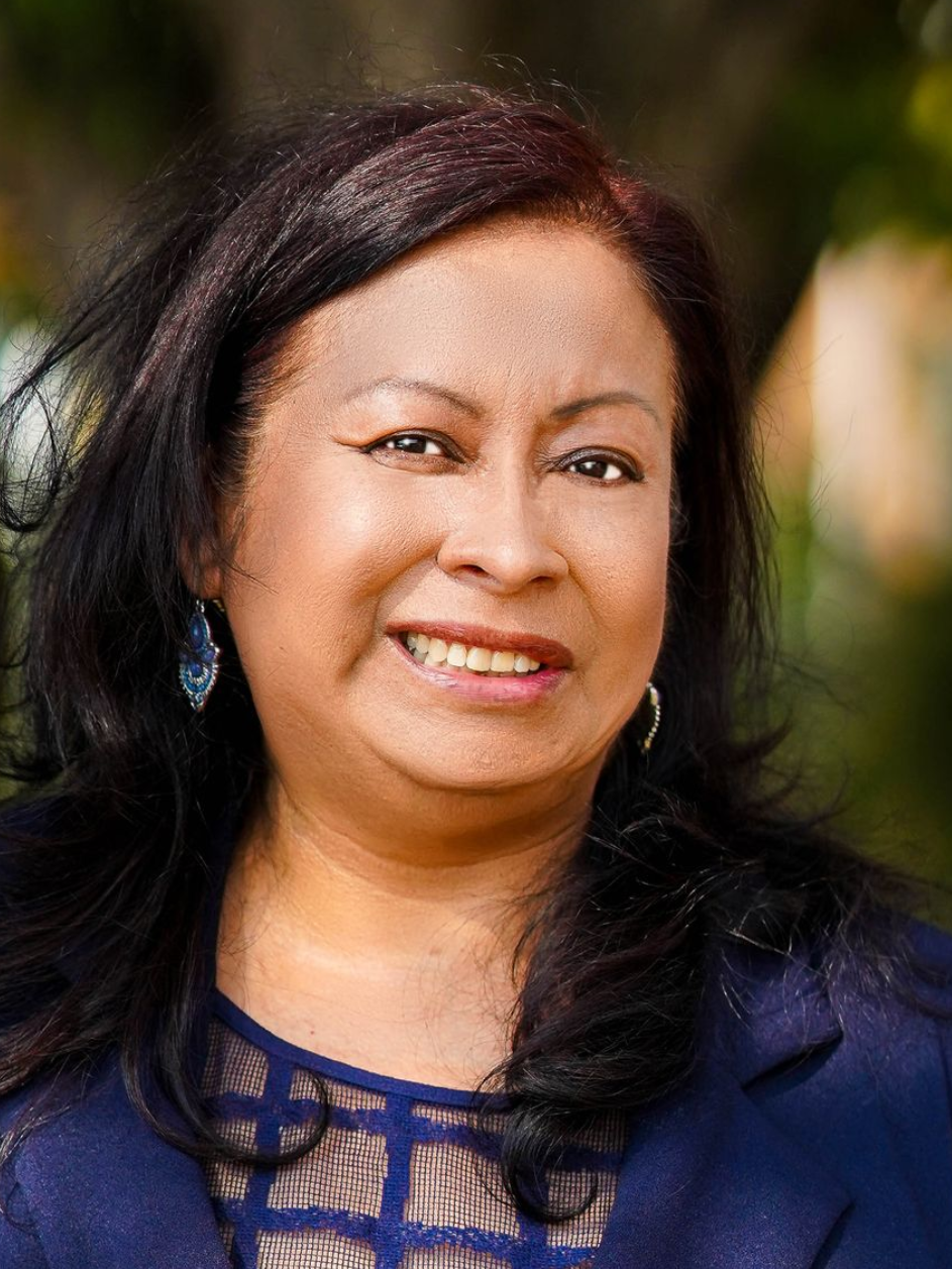 Woman with dark hair smiles, wearing a blue blazer over a patterned top, with a blurred outdoor background.