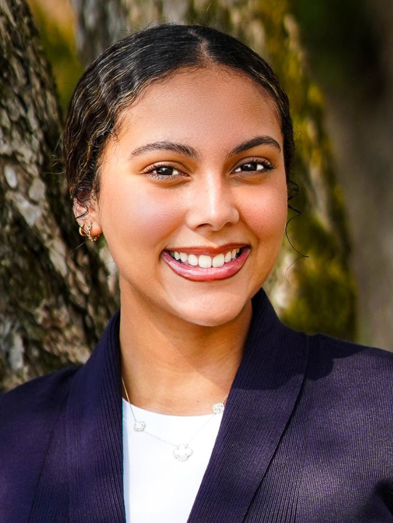 Woman with a wide smile, wearing a dark blazer and necklace, against a blurred tree background.