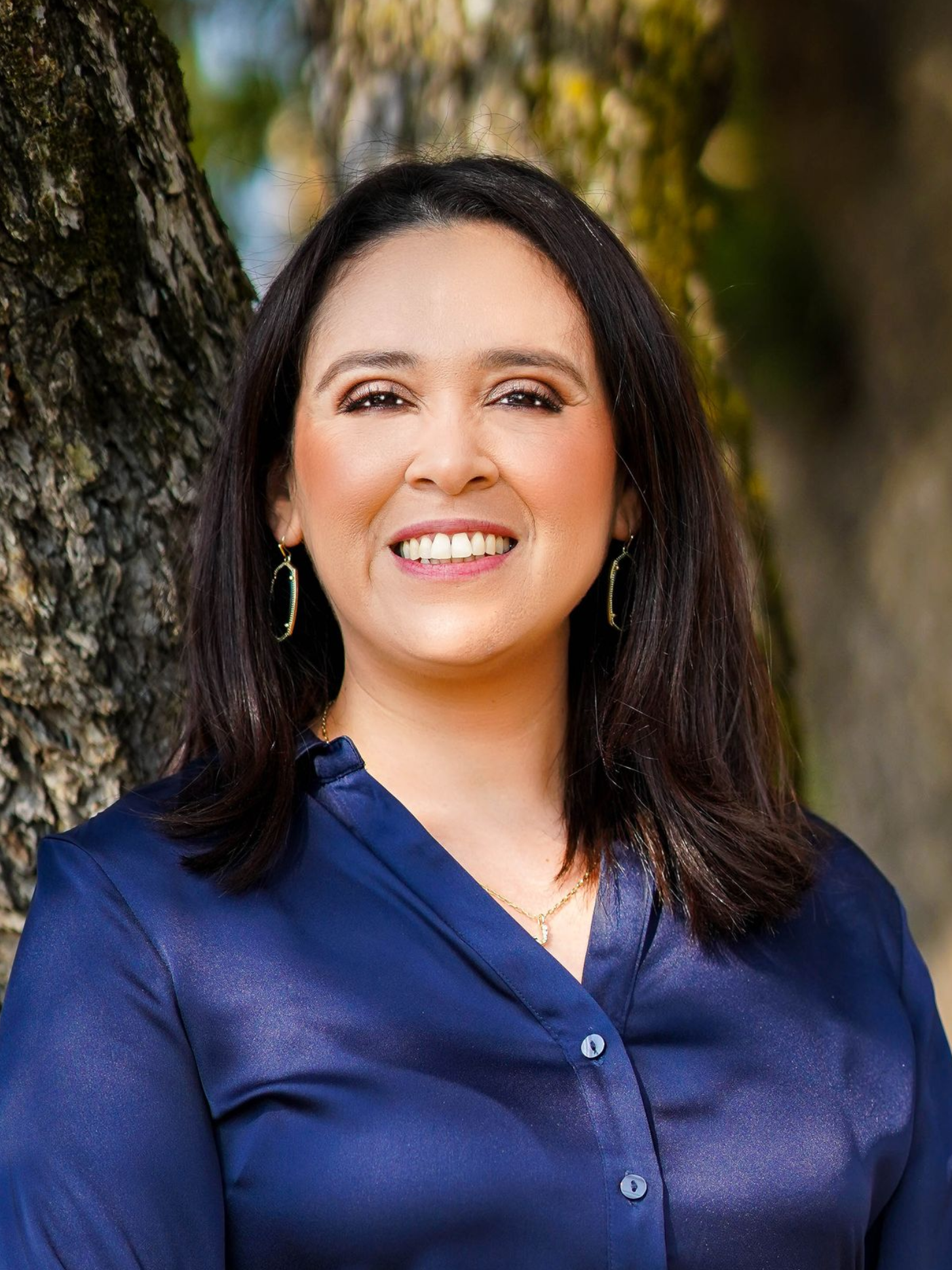 Woman with dark hair smiles, wearing a navy blue shirt, standing near a tree, outdoors.