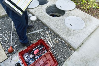 Worker beside open manhole with tools and red toolbox on gravel near concrete cover slabs