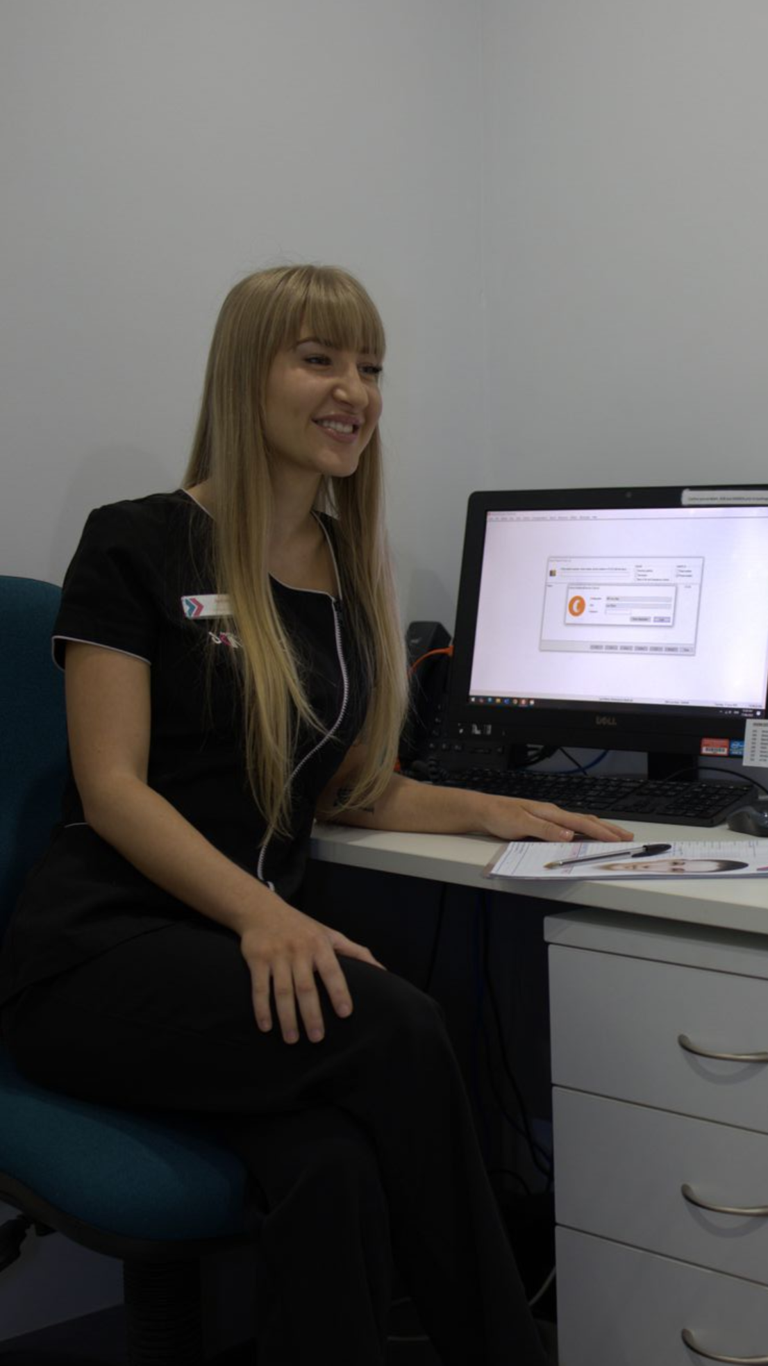 Woman seated at a desk beside a computer monitor, smiling in an office setting — Mackay Skin Clinic In North Mackay, QLD