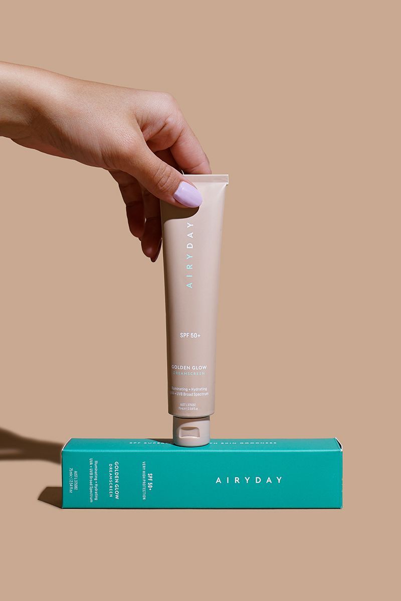 Hand holding a beige cosmetic tube above a turquoise box on a tan background