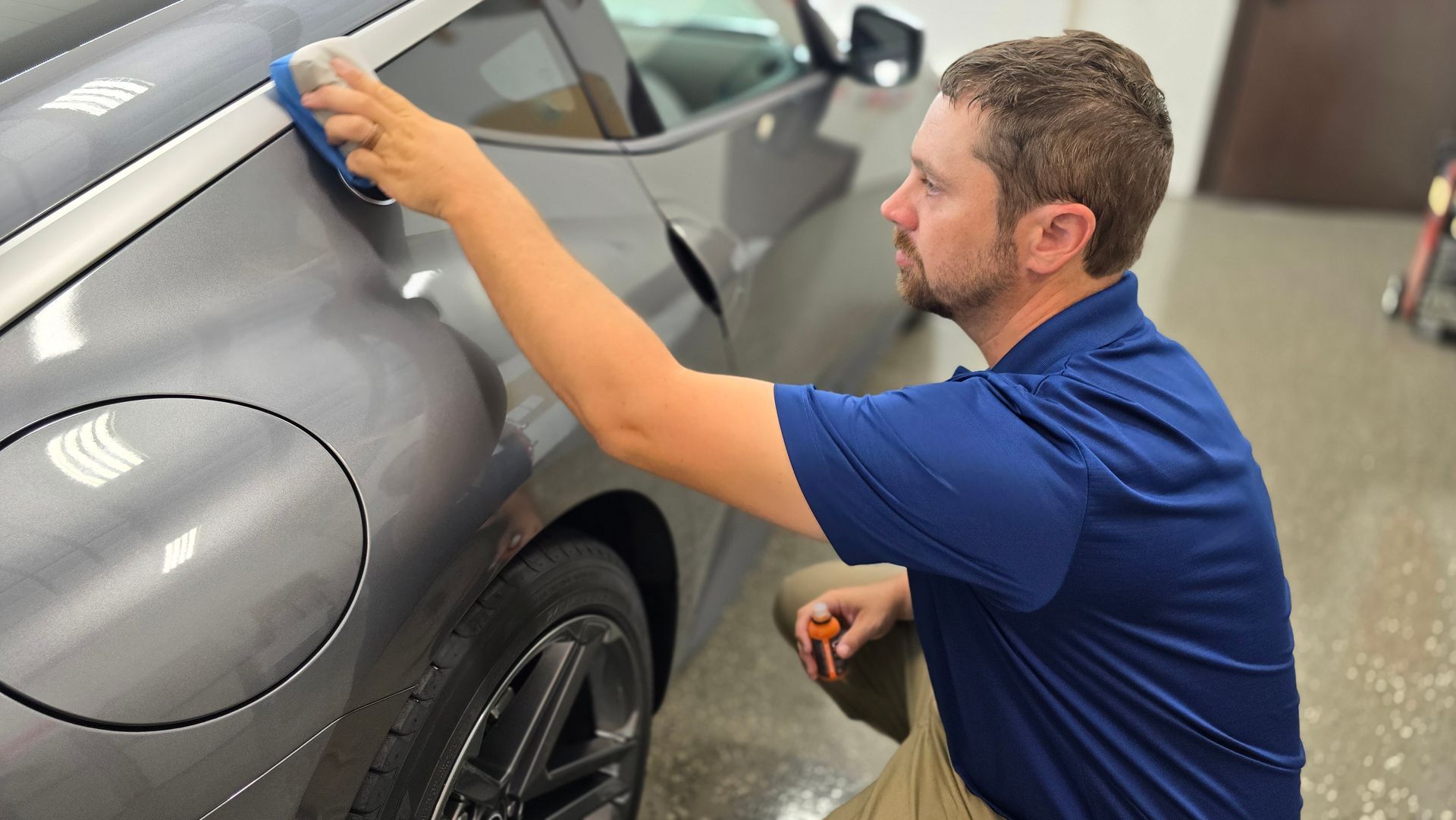 Man in blue shirt cleaning a gray car with a sponge in an indoor setting.