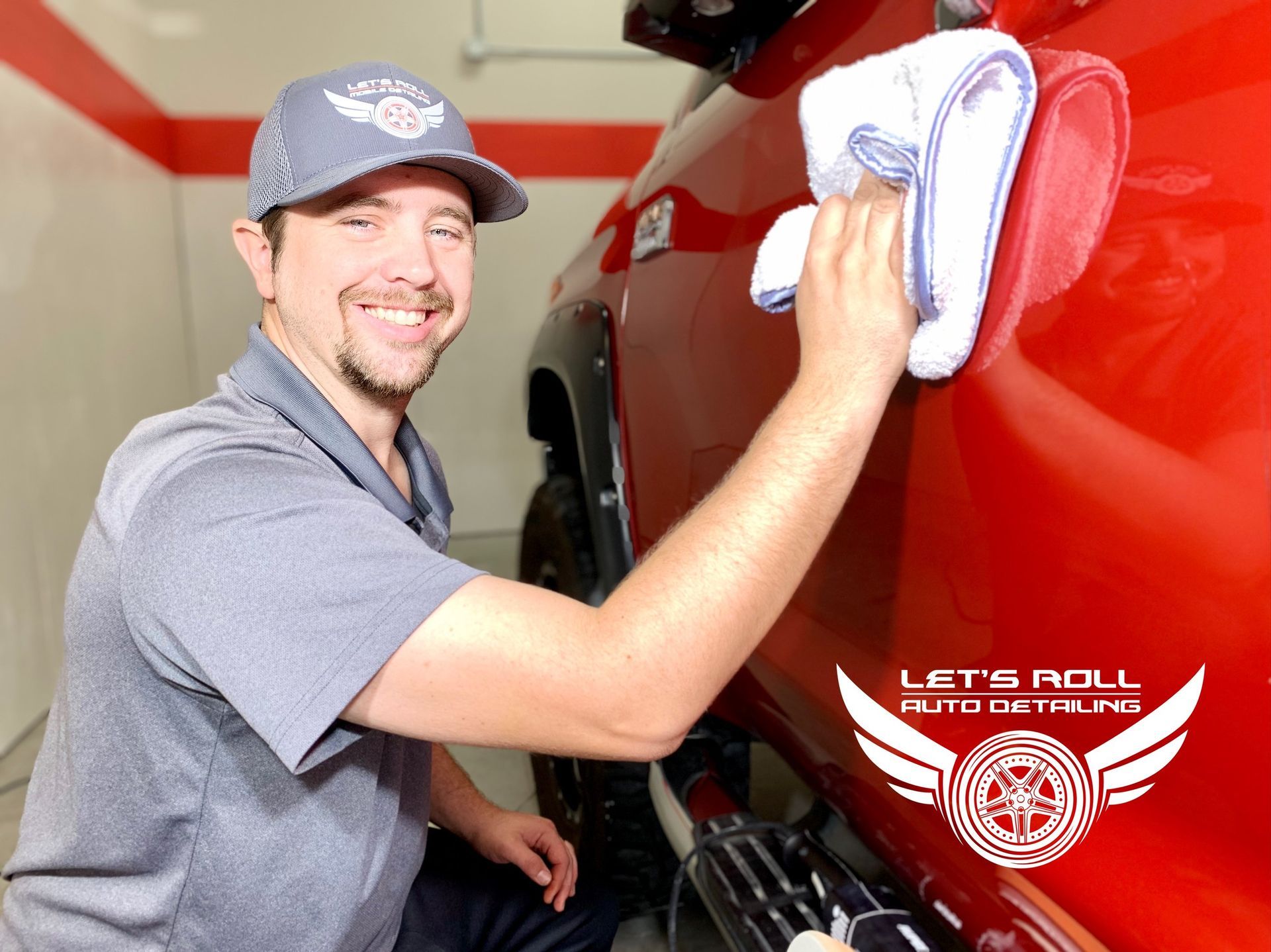 Man polishing a red car; wears a cap and smiles, in a garage with a 